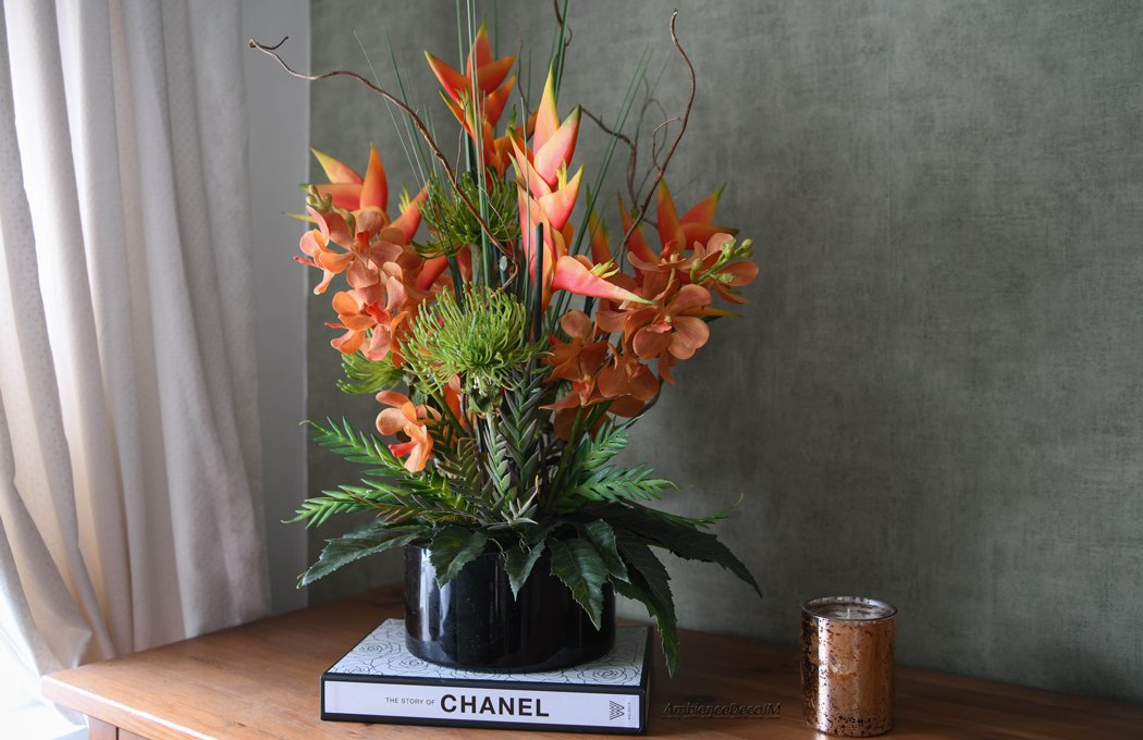 1cFauxTropicalCentrepieceLLW Luxury tropical artificial flower arrangement featuring vibrant orange heliconias, realistic orchid blooms, green pincushion protea flowers, architectural grasses, and decorative branches displayed in a sleek black vase. Premium faux floral centrepiece for modern and luxury interiors.