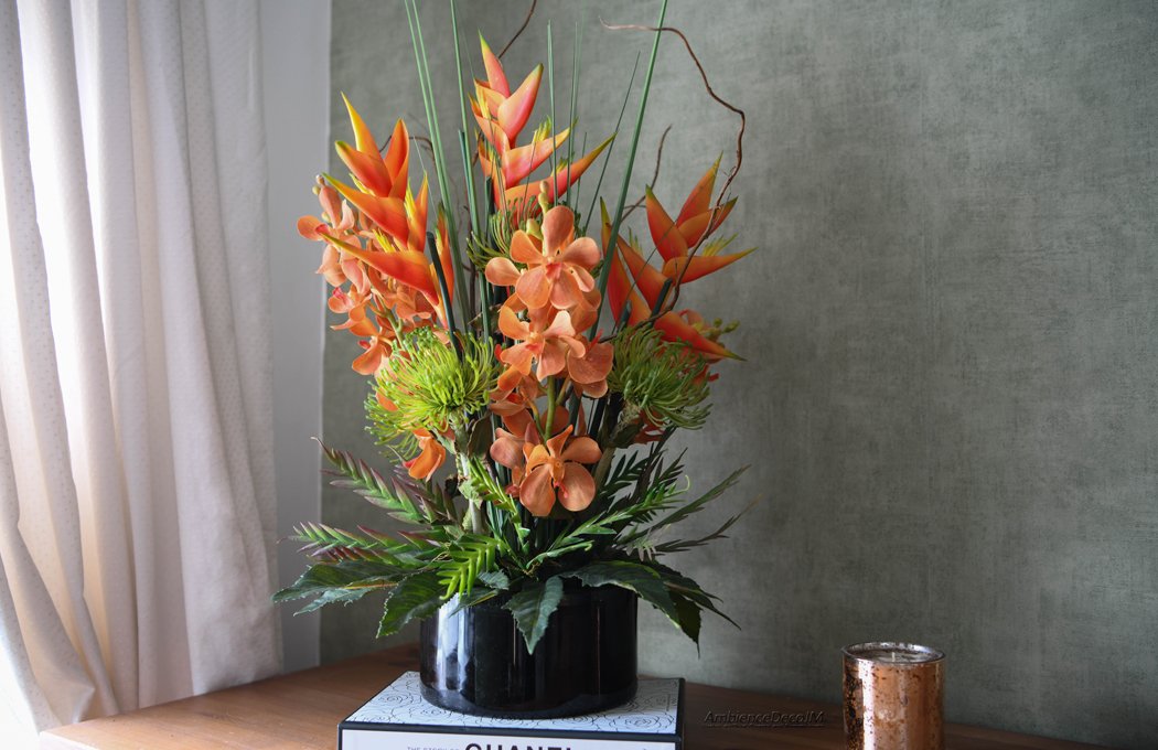 1fFauxTropicalCentrepieceLLW Luxury tropical artificial flower arrangement featuring vibrant orange heliconias, realistic orchid blooms, green pincushion protea flowers, architectural grasses, and decorative branches displayed in a sleek black vase. Premium faux floral centrepiece for modern and luxury interiors.