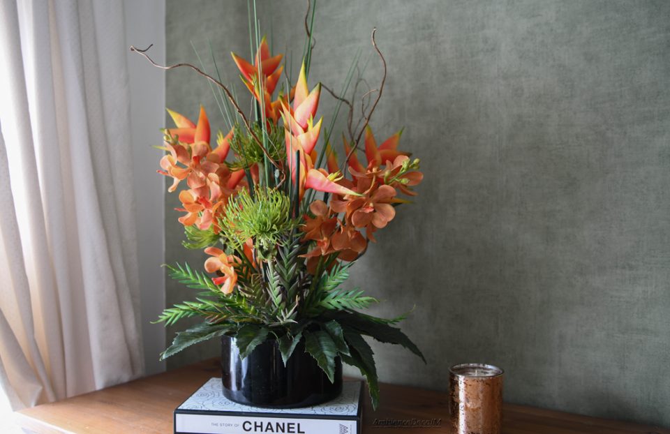 Luxury tropical artificial flower arrangement featuring vibrant orange heliconias, realistic orchid blooms, green pincushion protea flowers, architectural grasses, and decorative branches displayed in a sleek black vase. Premium faux floral centrepiece for modern and luxury interiors.