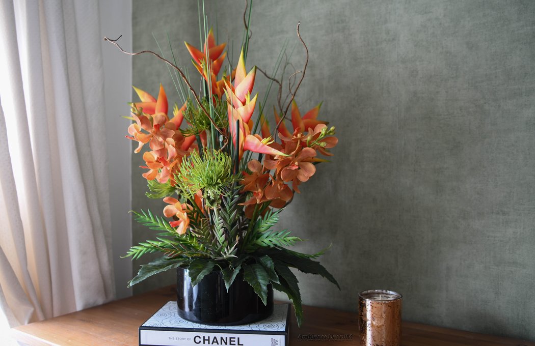 Luxury tropical artificial flower arrangement featuring vibrant orange heliconias, realistic orchid blooms, green pincushion protea flowers, architectural grasses, and decorative branches displayed in a sleek black vase. Premium faux floral centrepiece for modern and luxury interiors.